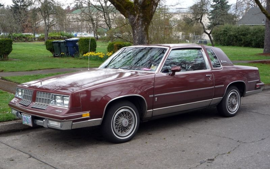 A maroon two-door vintage car is parked on a residential street beside a grassy park, with trees, flowers, and trash bins visible in the background.