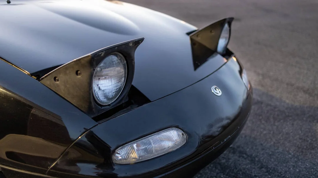 Close-up of the front end of a black car with pop-up headlights raised, showcasing one headlight clearly. The car is parked on a paved surface.