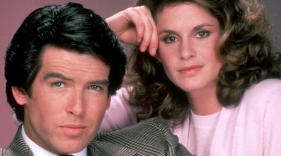 A man in a checkered suit and a woman in a light pink sweater pose closely together, both looking into the camera with neutral expressions against a pink background.