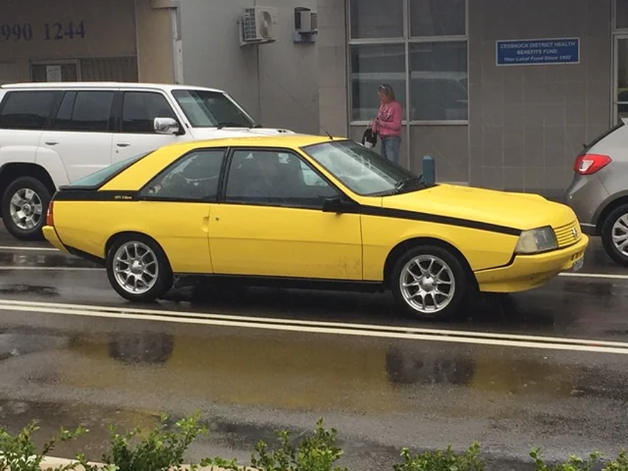 A yellow car with a unique, angular design is parked on a wet street beside other cars. A woman in a pink jacket walks on the sidewalk near a building in the background.