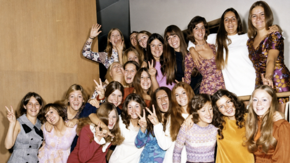 A group of young women, smiling and posing together in colorful 1970s-style clothing, make peace signs and laugh in a cheerful indoor setting.