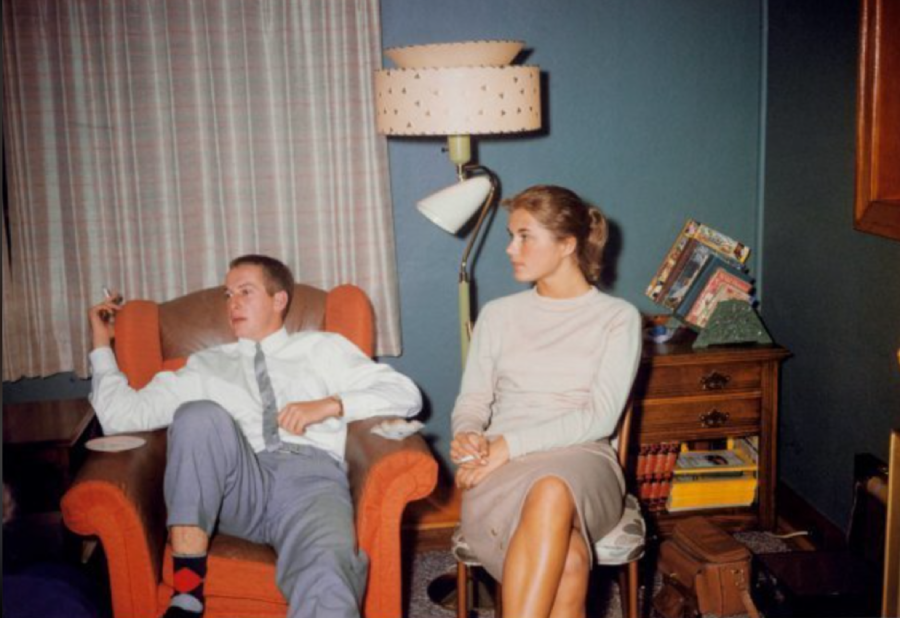 A man in a shirt and tie sits in an armchair, holding a cigarette, while a woman in a light sweater and skirt sits beside him. A lamp, books, and small table are in the background. The setting appears to be a living room.
