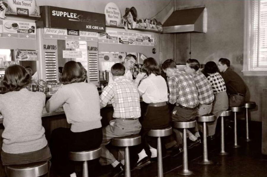 A group of teenagers sit on stools at a retro diner counter, facing away. They wear 1950s-style clothing. Behind the counter are soda fountains, a menu, and Coca-Cola signs.