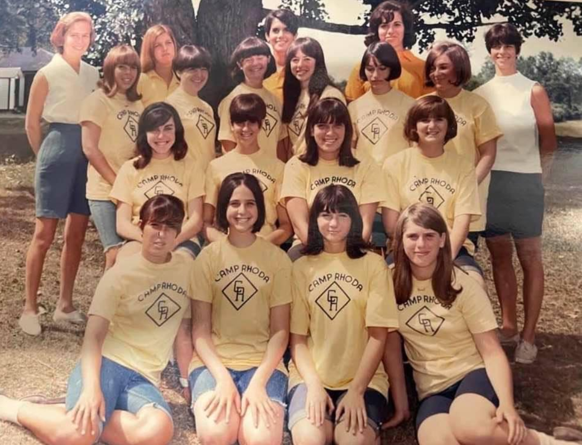 A group of teenage girls and two adult women pose outdoors under a tree, all wearing yellow "Camp Rhoad" T-shirts and shorts, smiling in a summer camp setting.