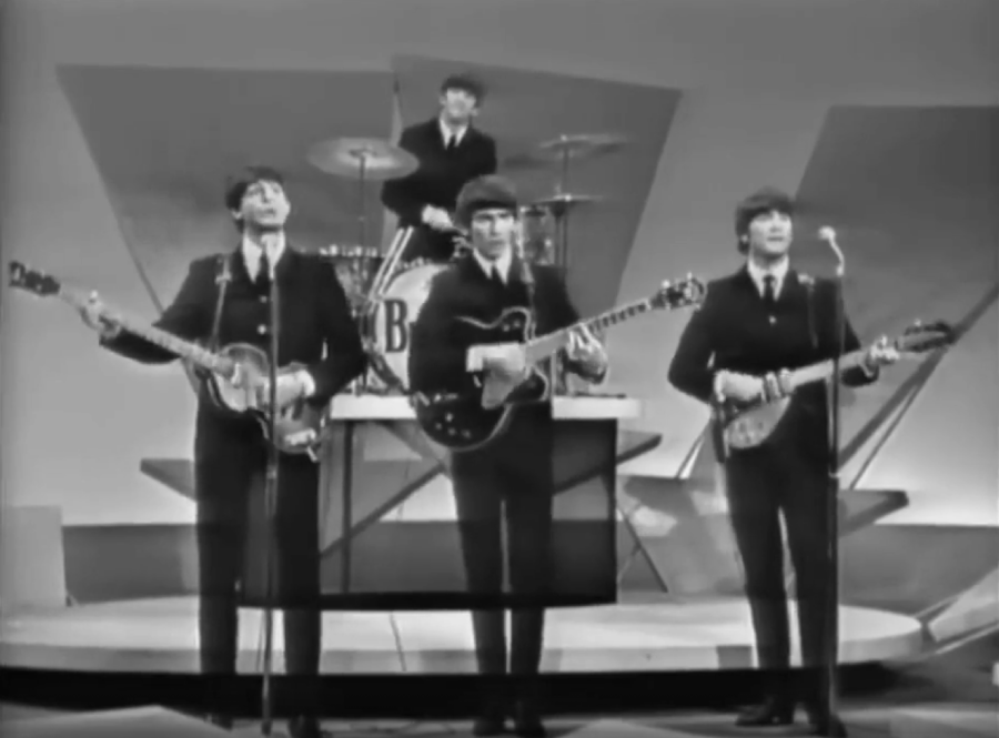 Four musicians in suits perform on a stage with guitars and drums. Three stand at the front with guitars, while one plays drums in the back. The stage has geometric shapes in the background.