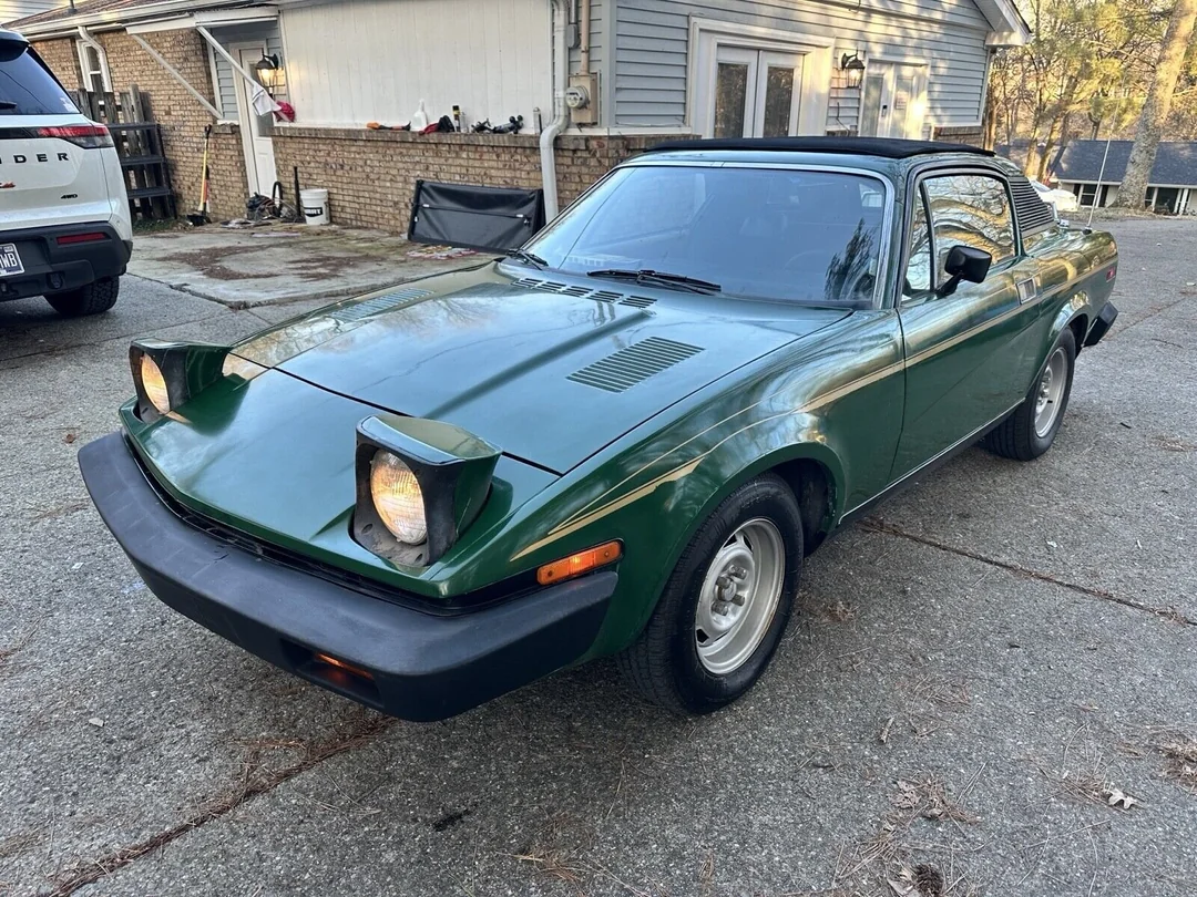 A green vintage sports car with pop-up headlights raised, parked on a concrete driveway in front of a house and garage, with other vehicles visible in the background.