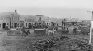 Historic black-and-white photo of a small frontier town&rsquo;s main street, featuring wooden buildings with business signs, horse-drawn wagons, people gathered, and logs piled in the foreground.