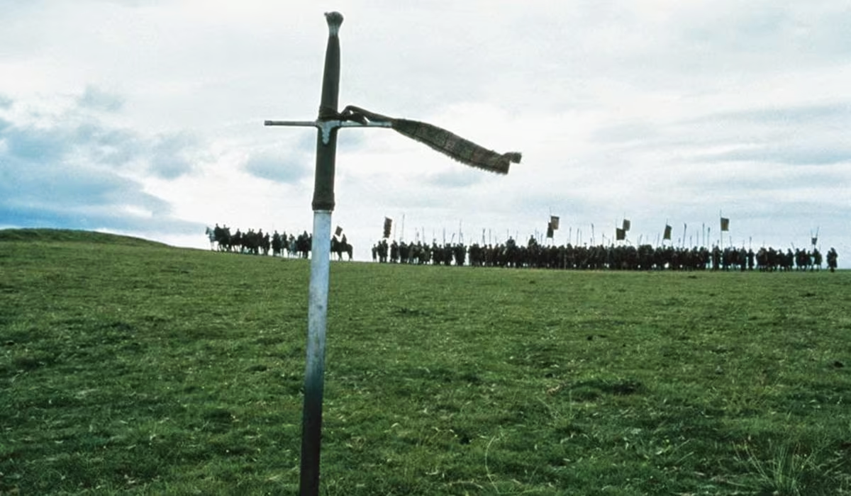 A large sword stands upright in a grassy field, with a tattered cloth tied to its hilt. In the background, a line of knights on horseback, holding banners, is silhouetted against a cloudy sky.
