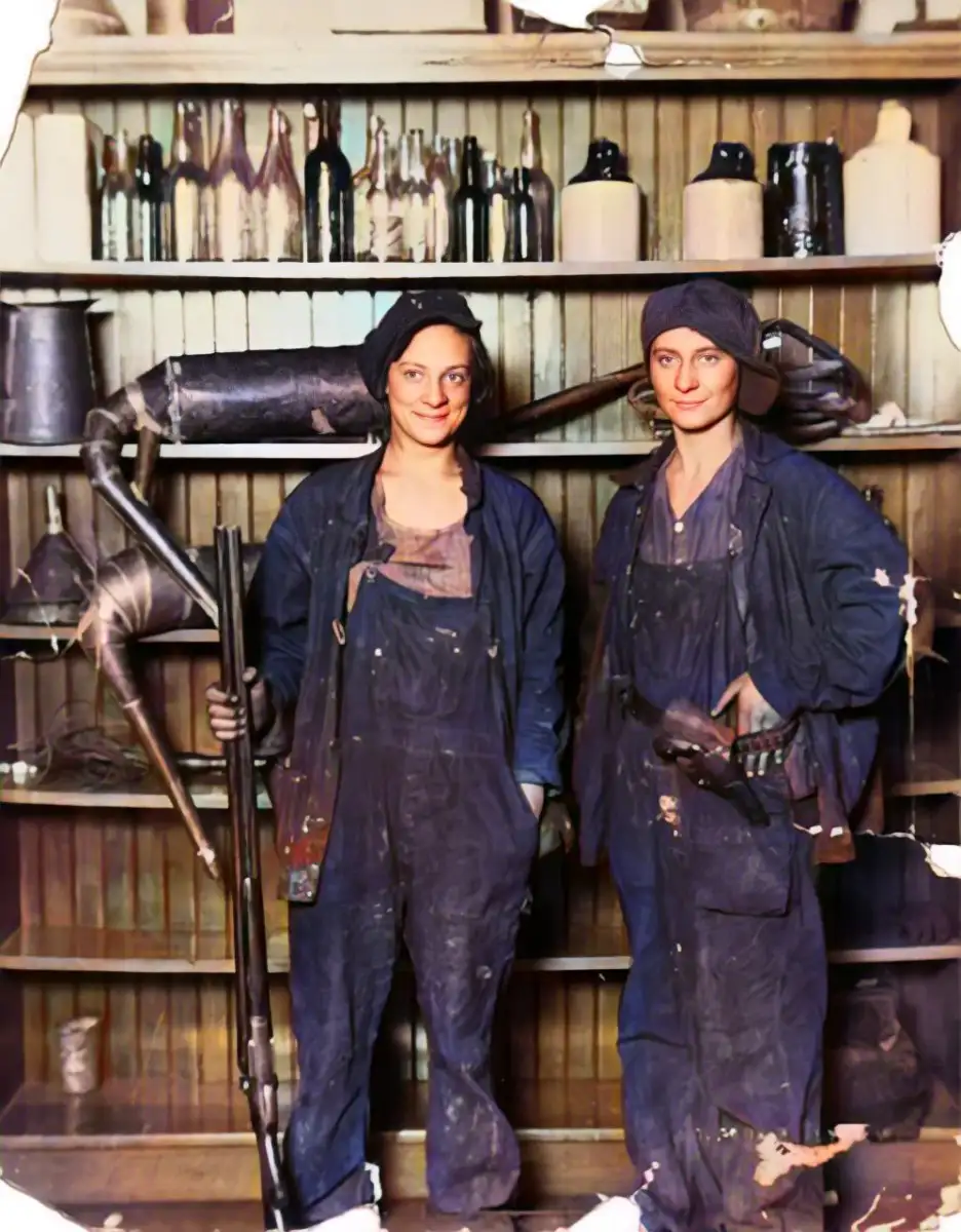 Two women in work overalls and headscarves stand in front of shelves with bottles and tools, holding industrial equipment. The photo appears vintage, with visible wear and discoloration at the edges.