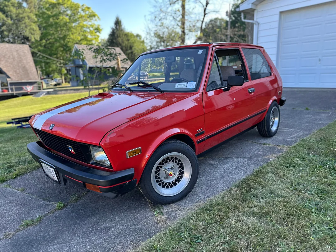 A shiny red vintage hatchback car is parked on a driveway beside a white garage, with green grass and trees in the background on a sunny day.