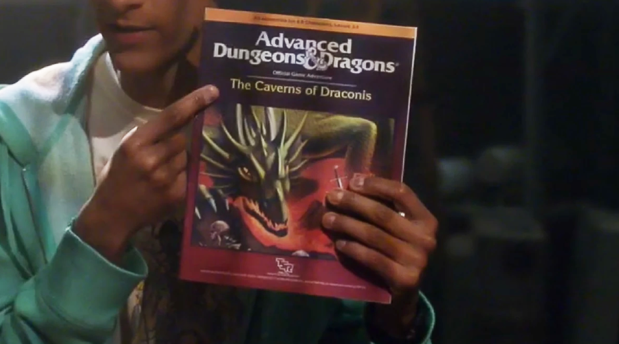 A person holds up and points to an "Advanced Dungeons & Dragons" game book titled "The Caverns of Draconis," featuring an illustration of a green dragon on the cover.