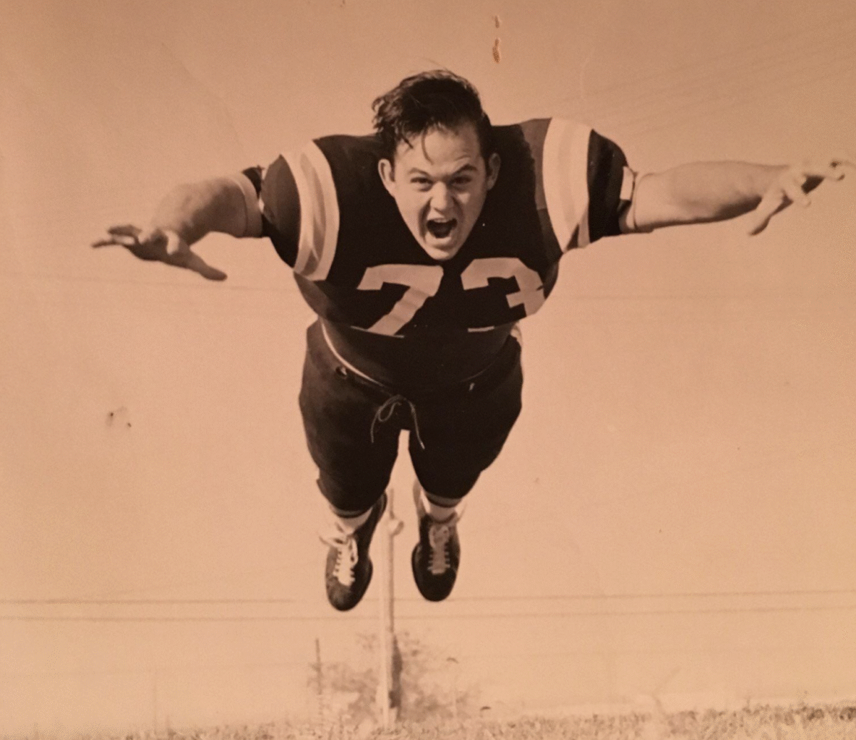 A vintage photo shows a football player wearing jersey number 73, captured mid-air with arms outstretched and a determined expression, as if diving or leaping forward. The image is sepia-toned.