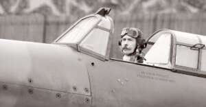 A pilot in vintage attire with goggles and a headset sits in the cockpit of an old airplane, looking ahead. The image is in black and white, emphasizing the historical atmosphere.