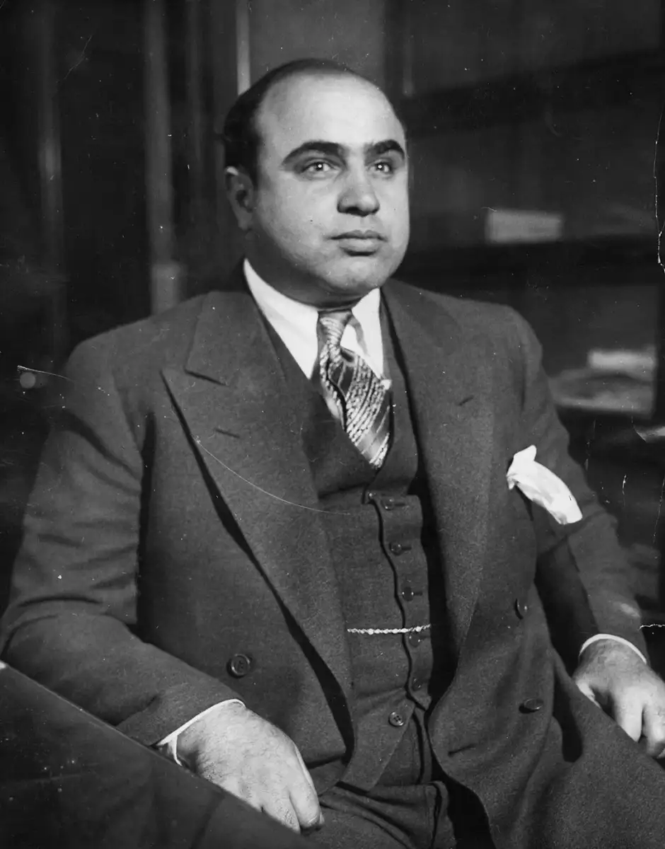 A man in a formal three-piece suit with a patterned tie and a pocket square sits at a desk, looking slightly to the side in a black-and-white photograph.
