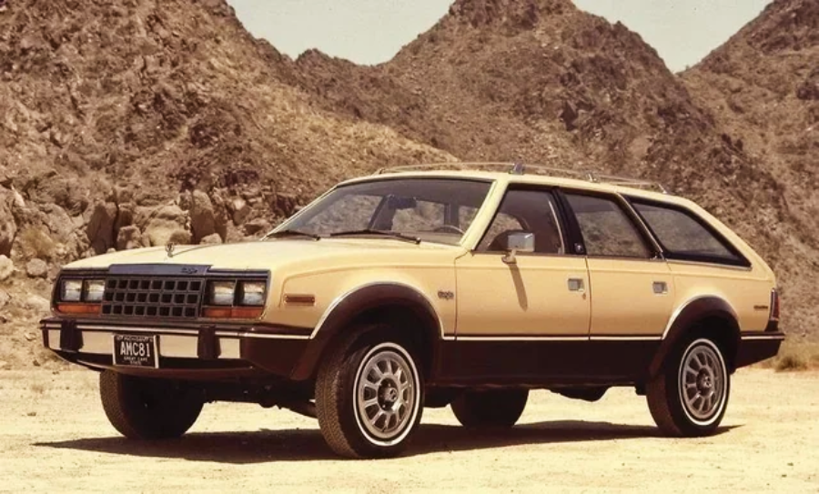 A tan and brown vintage station wagon is parked on sandy ground with rocky, arid mountains in the background under a clear sky.
