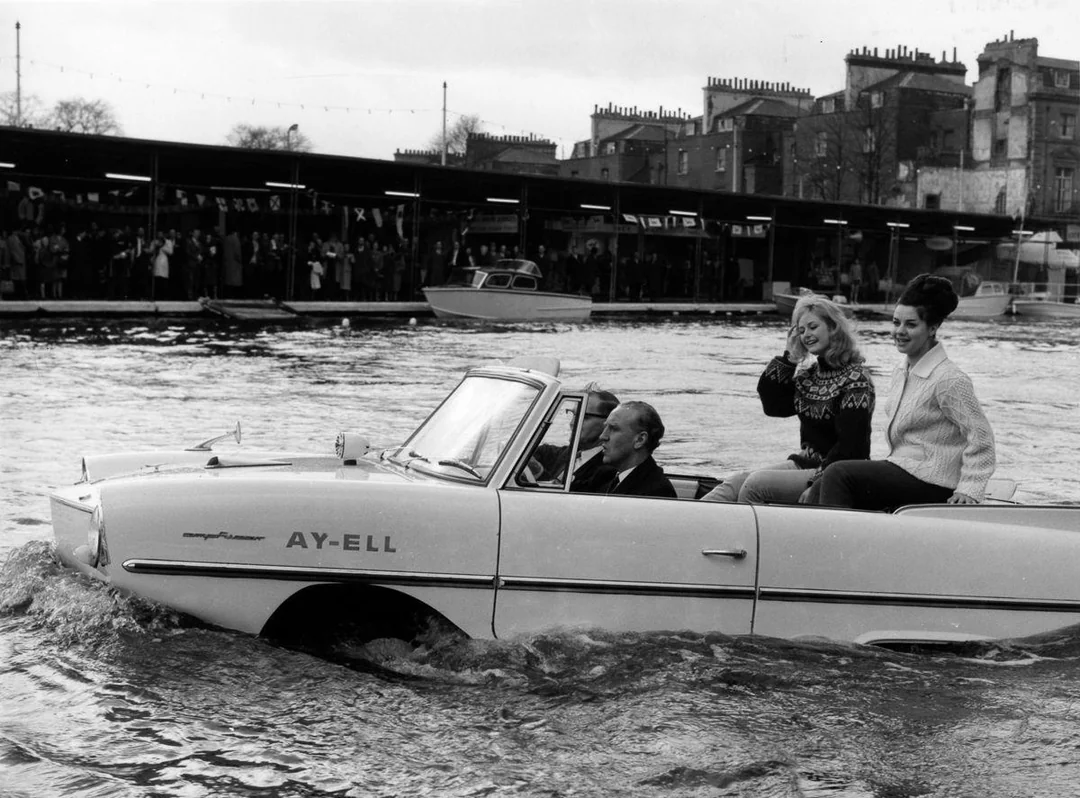A vintage amphibious car with three people inside drives through water. Two women sit in the back, one waving, as a man drives. Onlookers and buildings are visible along the distant waterfront.