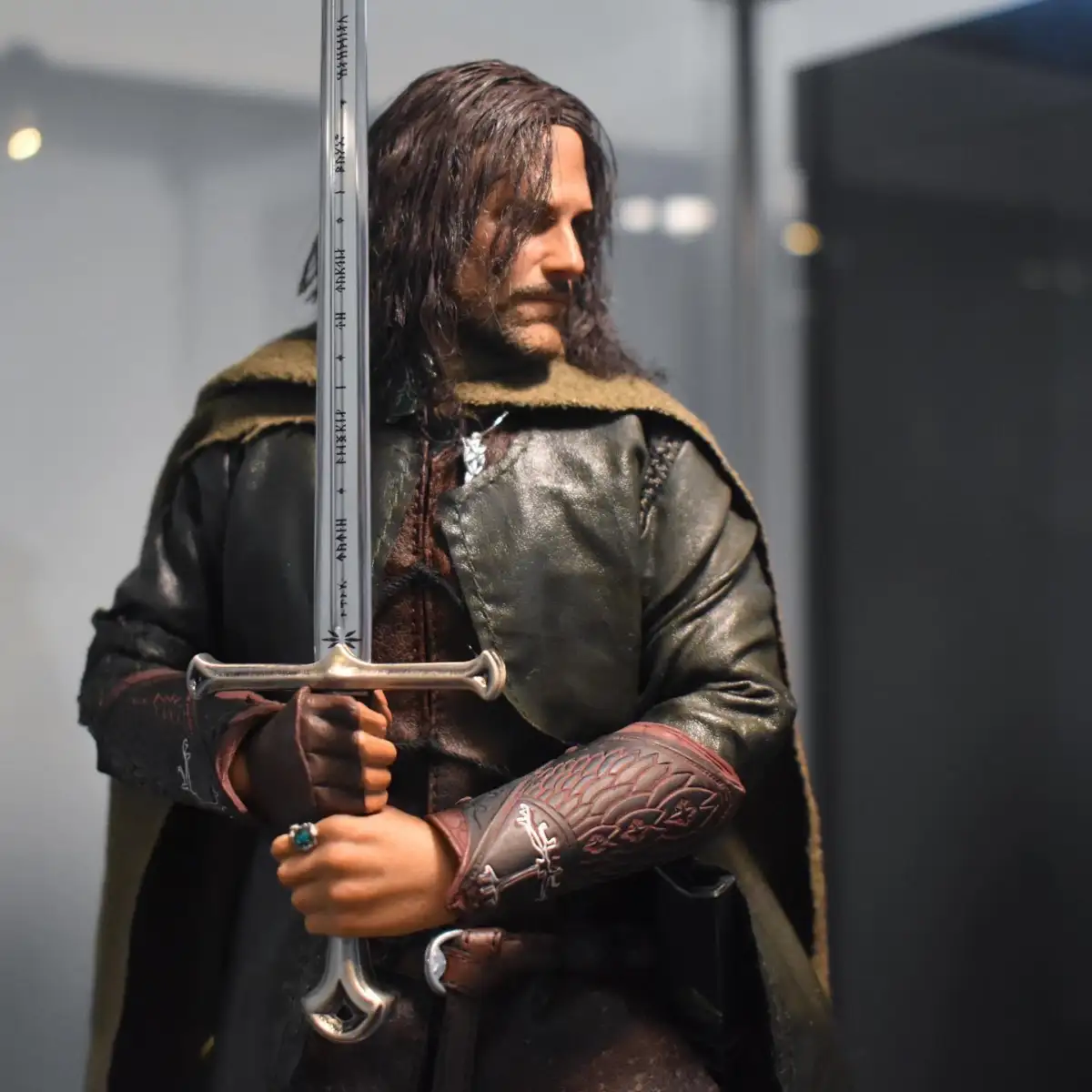 A figurine of a long-haired man in medieval clothing holds a large sword with both hands, wearing a green cloak and leather armor, looking to the side with a serious expression.