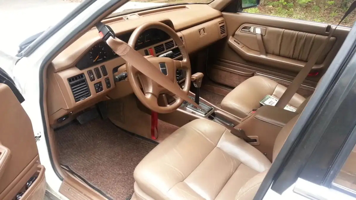 The interior of a vintage car with brown leather seats, brown dashboard, and a unique seatbelt design extending across the passenger seat to the B-pillar. The driver's door is open, revealing the detailed controls and trim.