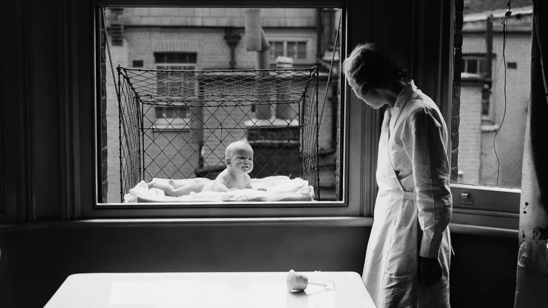 A woman looks out a window at a baby lying in a wire mesh crib suspended outside the building, with blankets beneath the baby and brick buildings visible in the background.