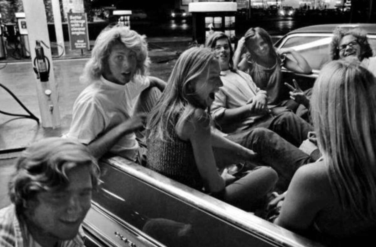 A group of young people laughing and talking while sitting in the back of a car at a gas station at night, illuminated by overhead lights.