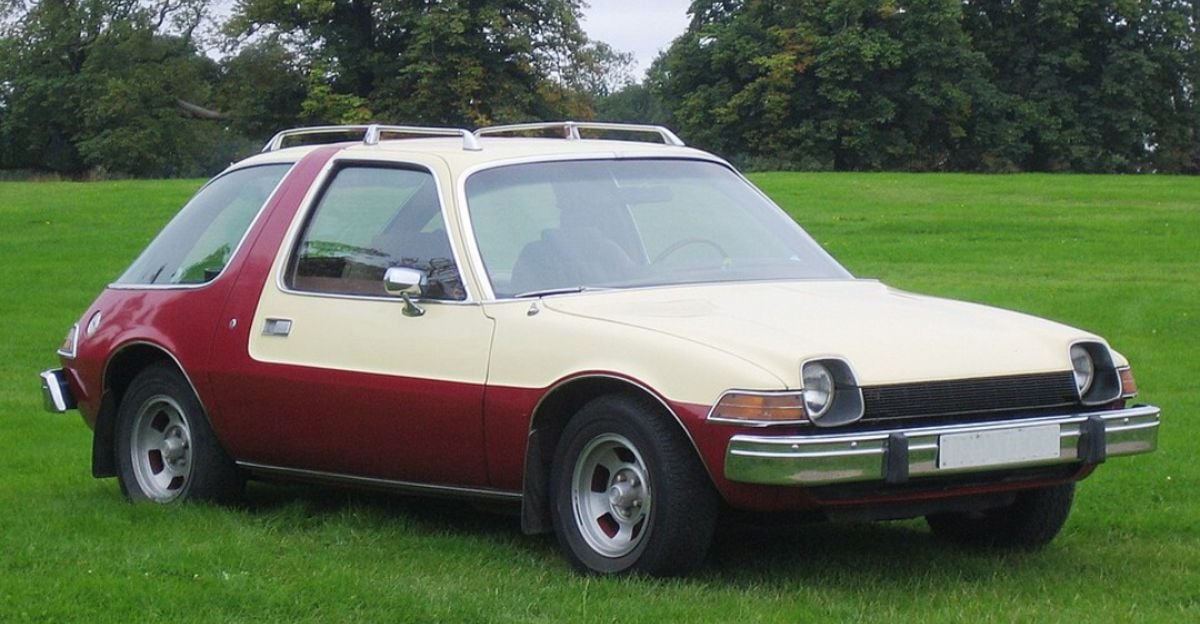 A classic two-tone car, cream on top and red on the bottom, is parked on a grassy field with trees in the background. The car has a rounded body, large windows, and chrome bumpers.