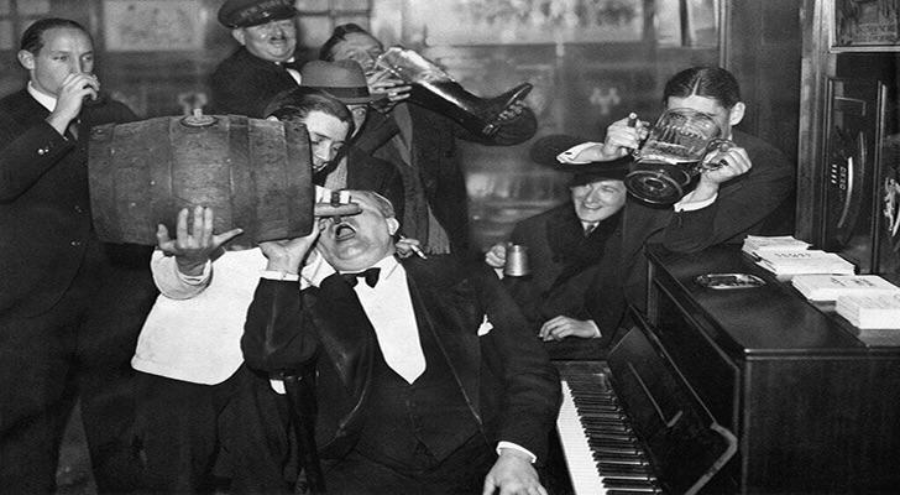 A group of people in formal attire enthusiastically drink alcohol from various containers, including a large barrel and a shoe, while gathered around a piano in what appears to be a lively, vintage party scene.