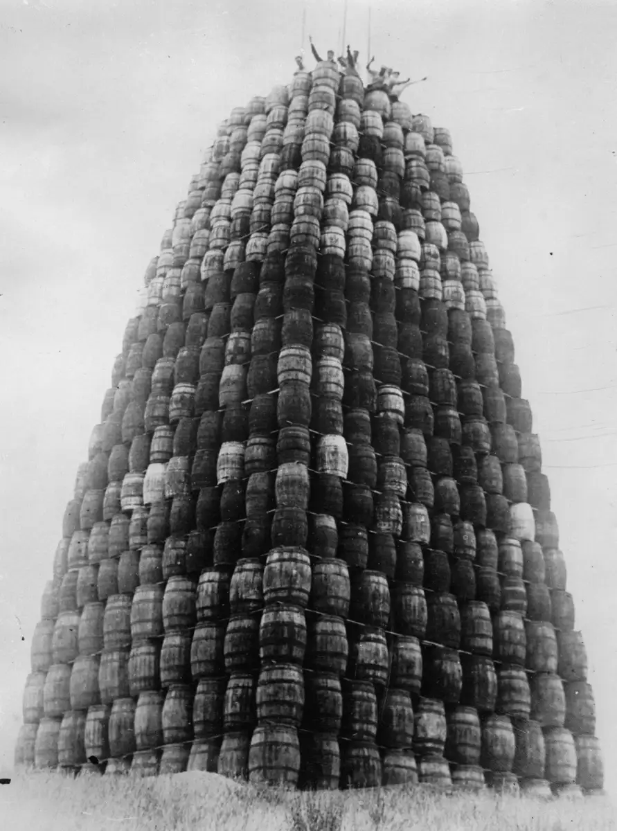 A large, tall pyramid made of stacked barrels stands outdoors, with several people visible on top, waving their arms. The photo is black and white and the sky is overcast.