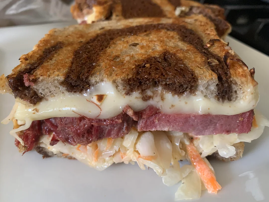 A close-up of a Reuben sandwich on marble rye bread, filled with melted Swiss cheese, corned beef, and coleslaw, served on a white plate.