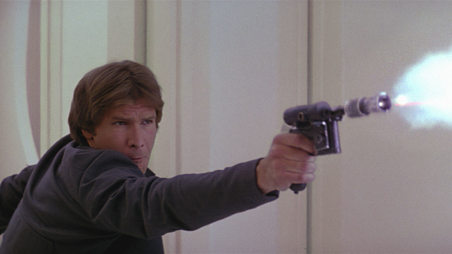 A man with brown hair and a serious expression aims and fires a blaster, creating a bright flash, in a futuristic, light-colored setting.