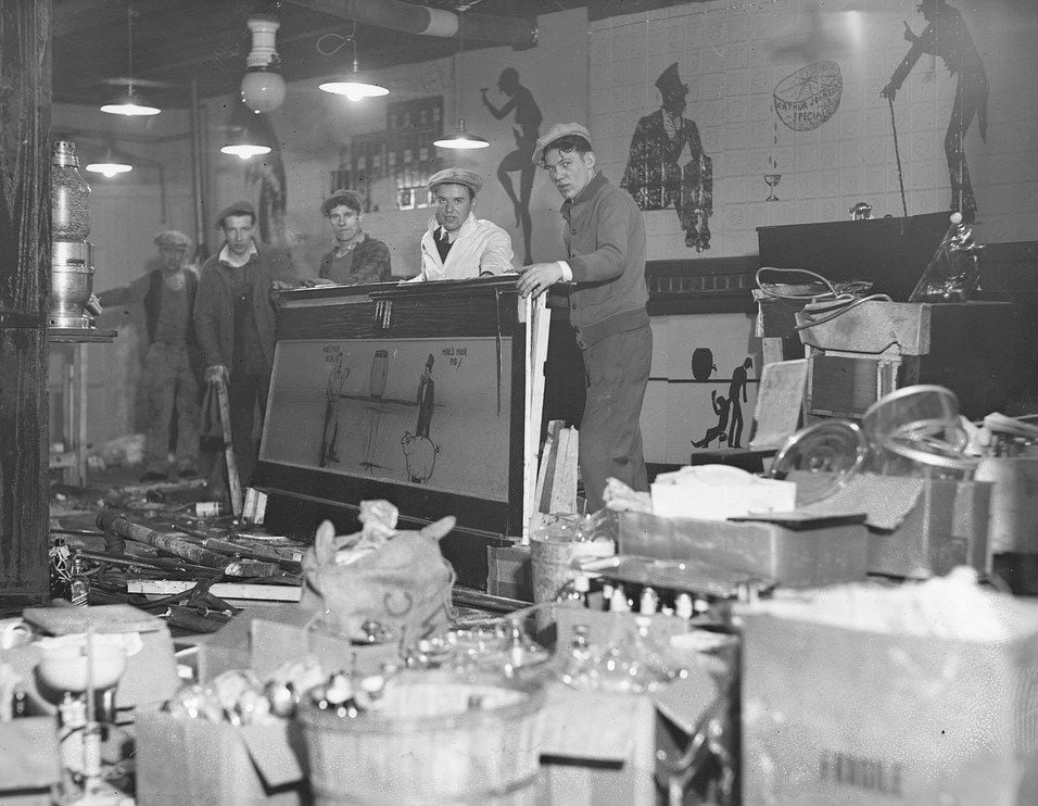 Five men stand behind a bar in a cluttered room filled with various confiscated liquor bottles and bar equipment, likely during the Prohibition era. The walls have painted silhouettes and there are overhead lights.