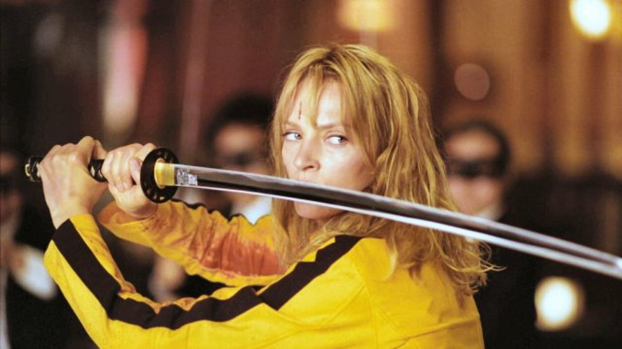 A woman in a yellow jacket holds a katana sword in a defensive stance, looking intensely to her right. The background is slightly blurred with people visible.