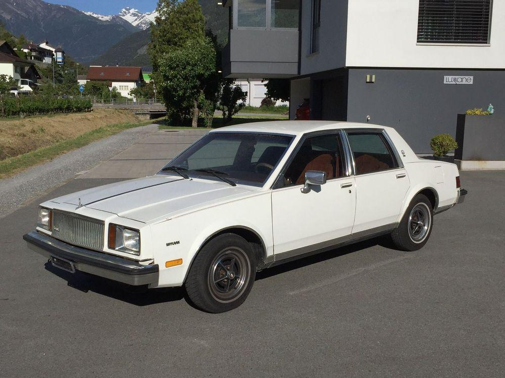 A white, vintage four-door sedan is parked on a paved driveway in front of a modern house, with mountains and greenery visible in the background.