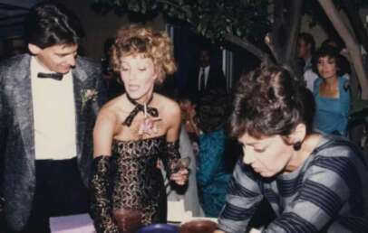 A man in a shiny suit and a woman in an off-the-shoulder patterned dress stand next to a woman in a striped dress who is bent over a table at a lively indoor event. Other people are visible in the background.