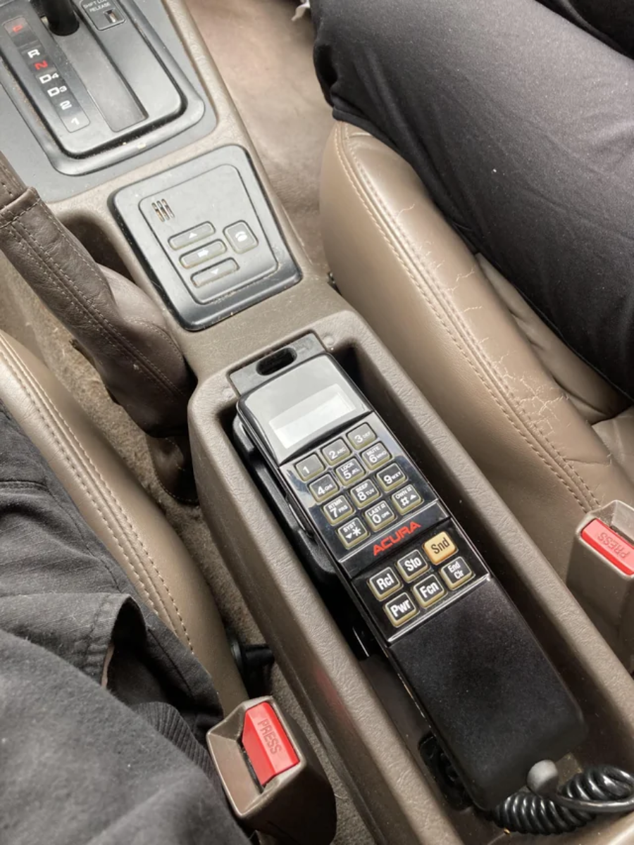 A vintage Acura car phone with a cord is mounted in the center console between two front seats, surrounded by seatbelt buckles and buttons for seat heating.