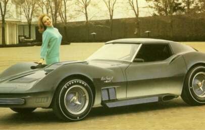 A woman in a turquoise dress leans against a sleek, futuristic silver sports car parked outdoors on a paved surface with trees and a building in the background.