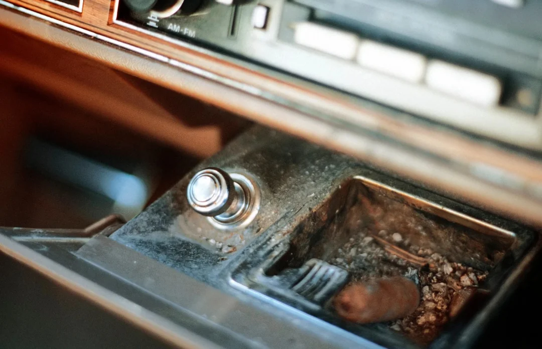 A close-up of an old car’s dashboard shows a cigarette lighter and a dirty ashtray with ashes and a used cigarette inside. The surrounding area appears dusty and worn.