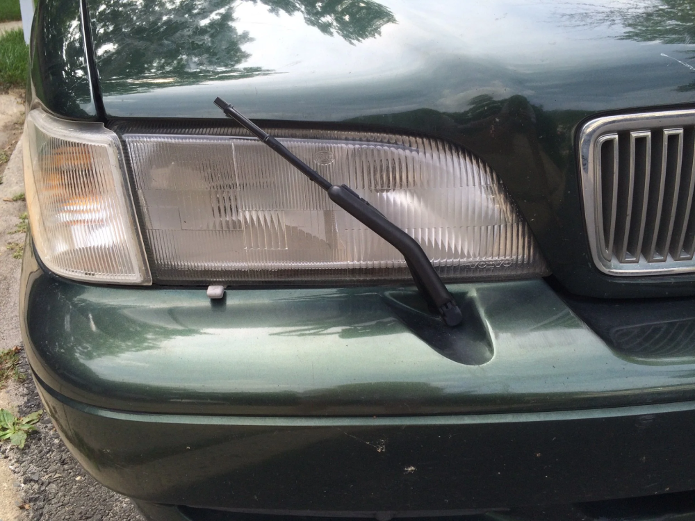 Close-up of a green car’s front headlight with a small windshield wiper mounted on the headlight, reflecting nearby trees and sky.