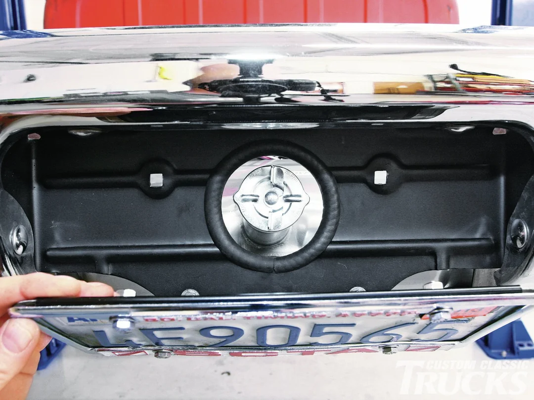 A close-up of a vintage vehicle’s rear, showing a license plate hinged open to reveal the gas cap behind it. A hand is holding the plate down, exposing the fuel filler area beneath chrome trim.
