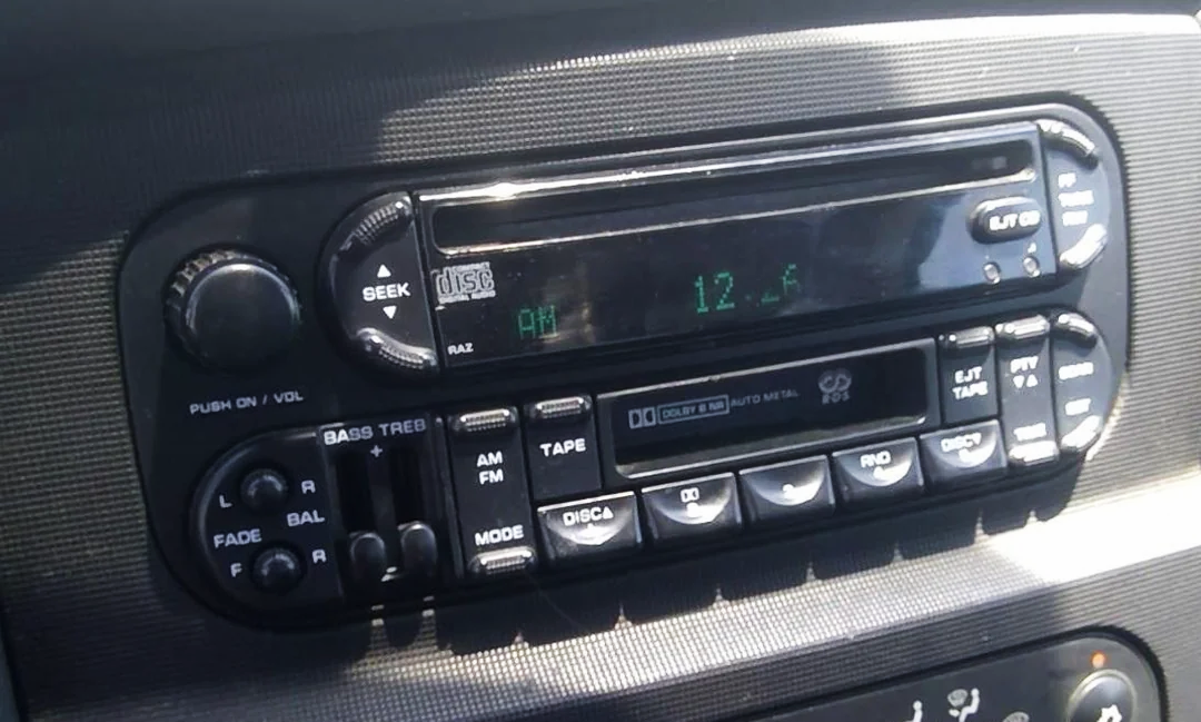 Close-up of a car stereo system displaying "12:46" on the digital clock. The radio has buttons for AM/FM, tape, CD, volume, seek, and other audio controls. The surface appears slightly worn.