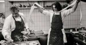 Two chefs in striped aprons work in a kitchen; one focuses on preparing food while the other holds up a long strip of pasta dough, stretching it wide with both hands. Shelves and kitchen equipment are visible in the background.