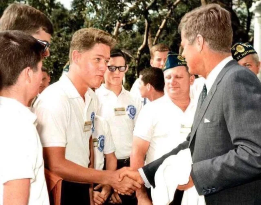 A young man in a white shirt shakes hands with an older man in a suit outdoors, surrounded by other young men in white shirts and hats, with trees visible in the background.