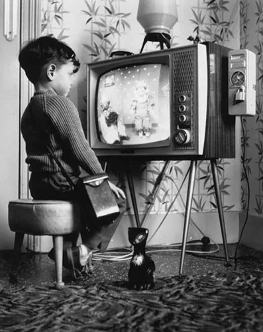 A young boy sits on a small chair, watching a vintage television. He holds a lunchbox and a ceramic cat figurine is on the floor beside him. The room has patterned wallpaper and a textured rug.