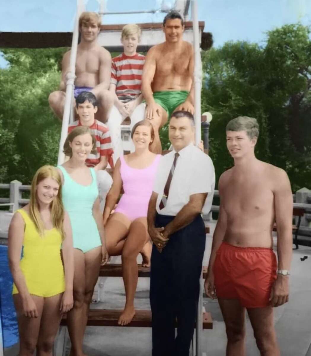 A group of people in swimsuits and one man in a shirt and tie pose by a poolside ladder on a sunny day, with trees and a fence in the background.