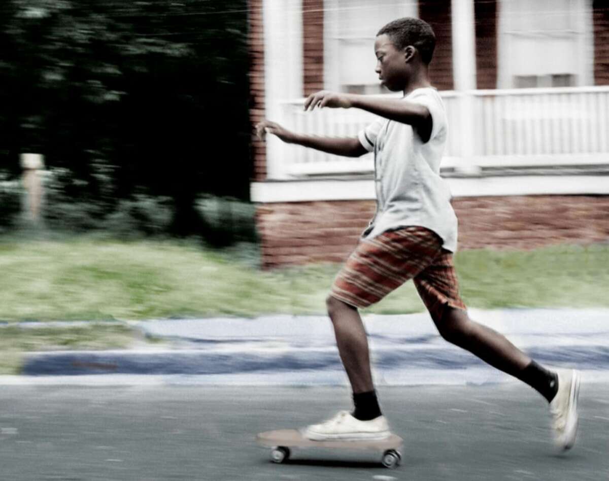 A boy wearing a gray shirt and plaid shorts rides a skateboard down a street, pushing off with one foot. The background is blurred, suggesting motion, and a red brick building with a white porch is visible behind him.