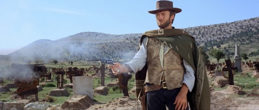 A man in a cowboy hat and poncho stands in a graveyard, holding a smoking revolver. He is surrounded by tombstones and dry, hilly terrain under a clear blue sky.