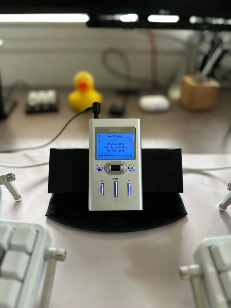 A silver MP3 player with a blue-lit screen sits on a desk stand between two white keyboards. A yellow rubber duck and other office items are blurred in the background.