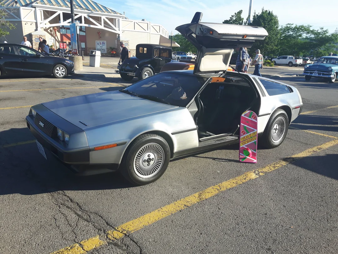 A silver DeLorean car with gull-wing doors open is parked in a lot. A pink hoverboard is propped against the car, referencing the "Back to the Future" movies. Other cars and people are visible in the background.