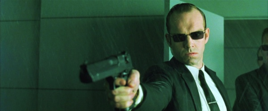 A man in a black suit and sunglasses points a gun forward with a serious expression, standing in a green-tinted, modern, indoor setting.