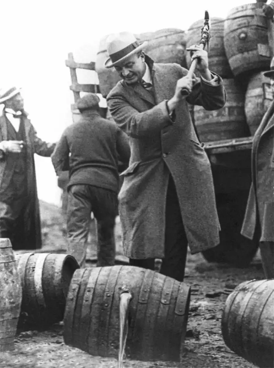 A man in a coat and hat uses a tool to smash a barrel, spilling liquid onto the ground, while others stand near a truck loaded with barrels in the background. The scene appears to be from the early 20th century.