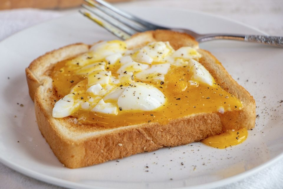 A slice of toasted bread topped with chopped soft-boiled eggs and runny yolk, sprinkled with black pepper, on a white plate with a fork in the background.